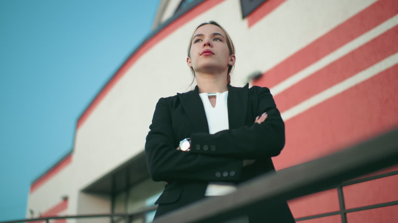 Young real estate manager in black blazer folds hands standing confidently in front of organization with red and white striped wall, security camera on building, leadership, and success