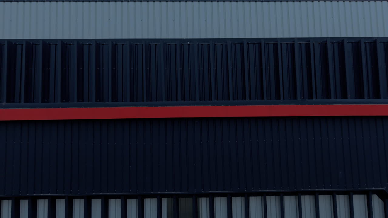 Panoramique aérien rapproché d'une installation de production moderne dotée de murs en tôle ondulée bleu foncé et gris avec une bande rouge vive.