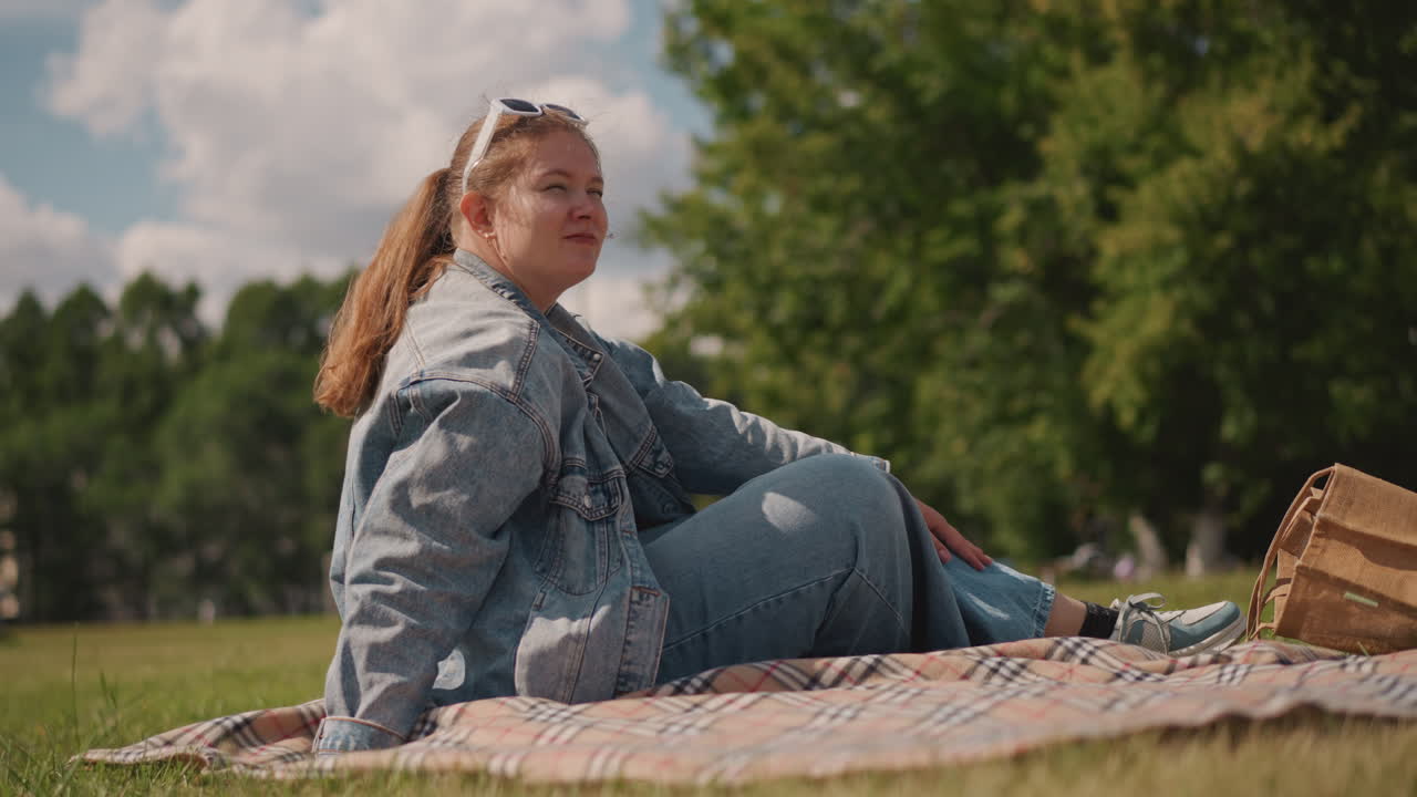 Kvinna njuter av utomhusrekreation. En dam sitter på en filt i solljuset. En kvinna kopplar av utomhus under en fridfull himmel. Damen sitter bekvämt på filten, omgiven av träd under en lugnande himmel.