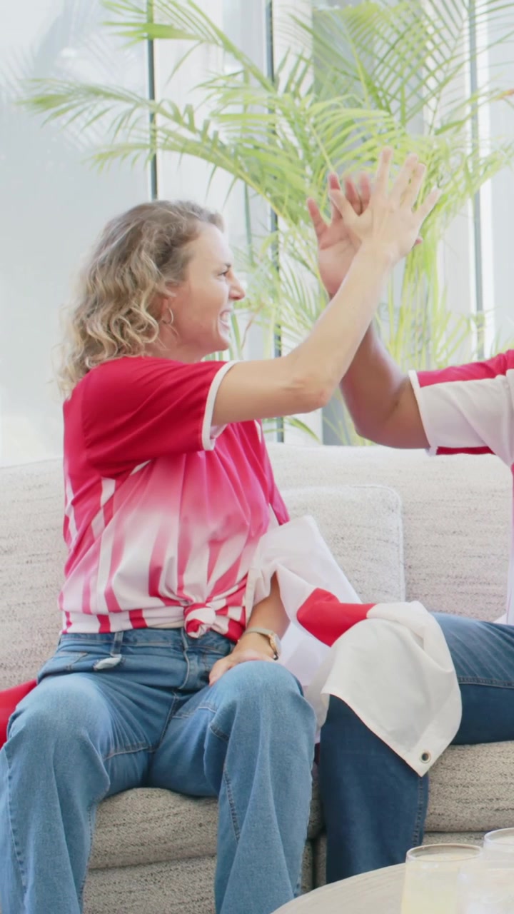 Vertical video: Cheering woman in red jersey turning to high-five man in red on couch after TV goal