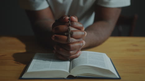 hands of man holding rosary beads with crucifix and praying with holy bible