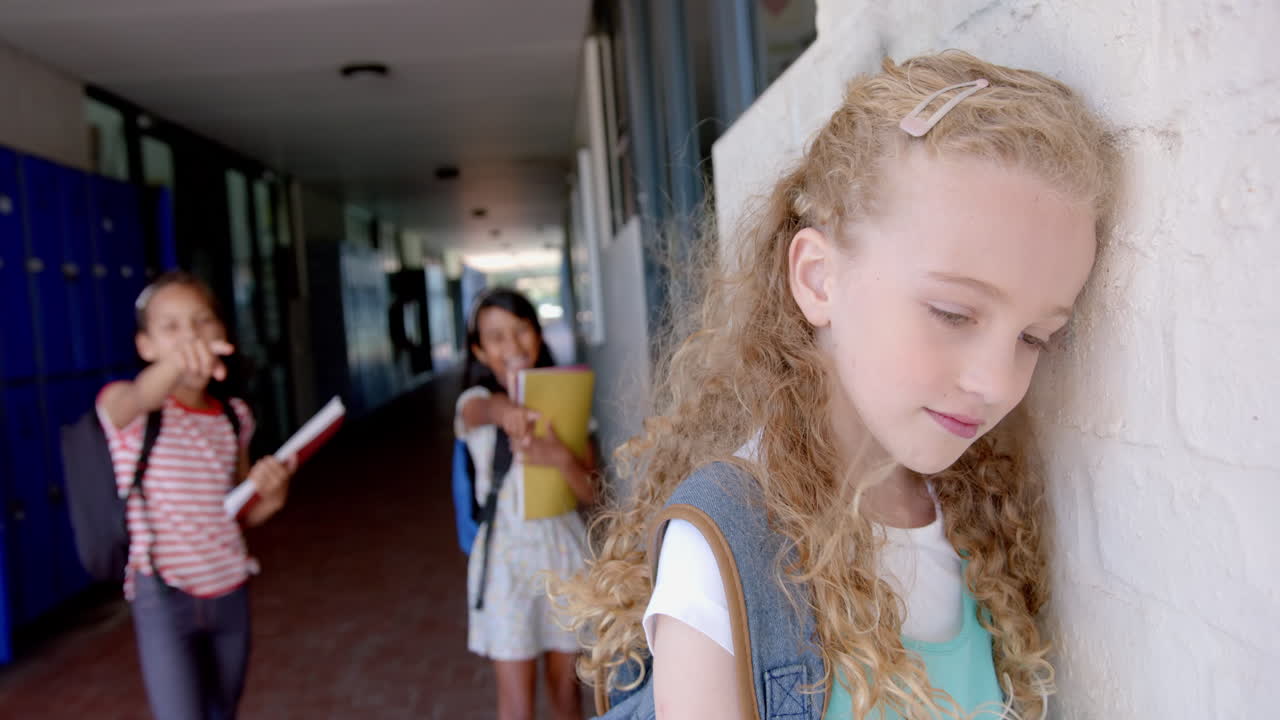 In school, sad girl leaning against wall while two students teasing her in hallway