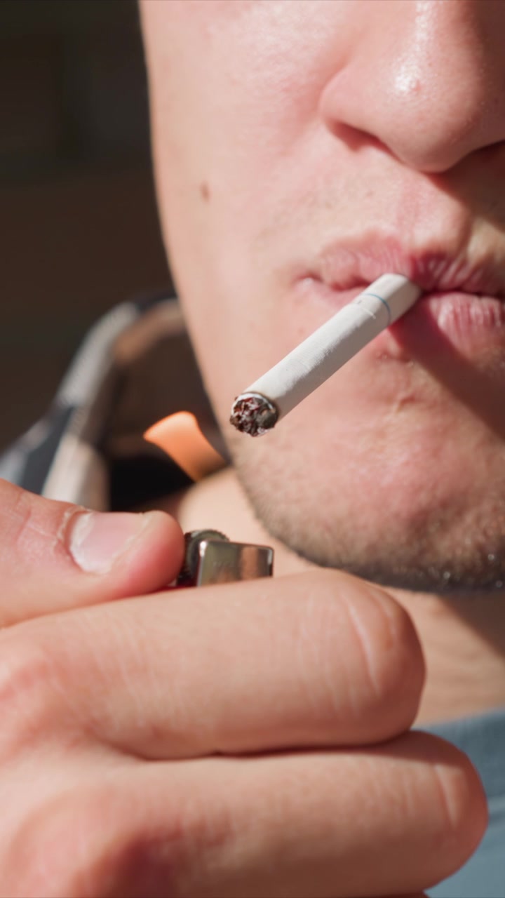 Primer plano de un hombre encendiendo y fumando un cigarrillo. La mano sostiene un encendedor mientras lo acerca para prender el extremo sin encender, con humo visible de uso anterior. La luz del sol crea sombras suaves en la piel.
