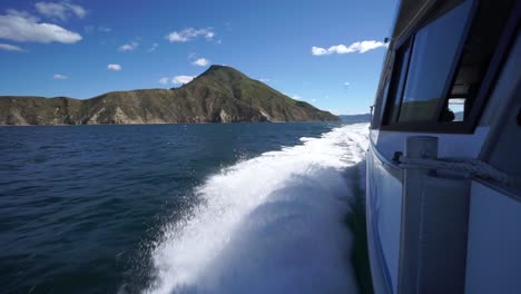 Foamy-waves-breaking-on-side-of-cruise-boat-in-Marlborough-Sounds,-New-Zealand-with-green-hills-in-background