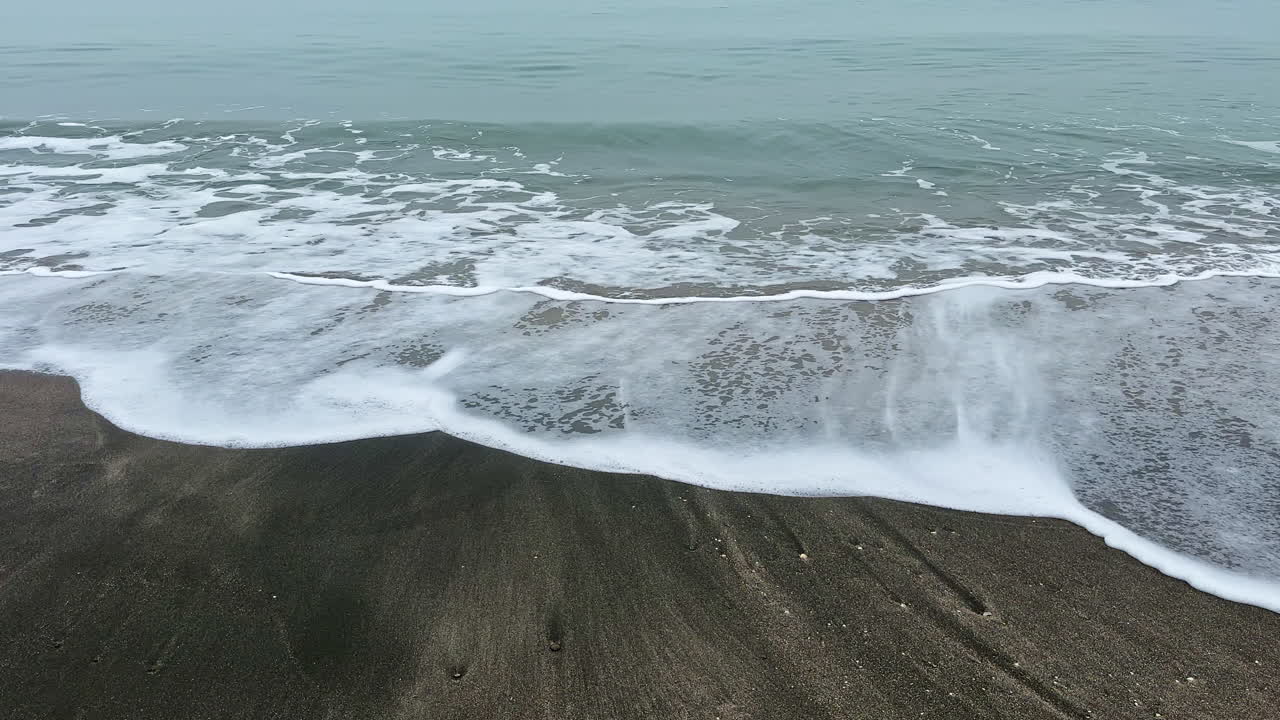 Le onde si infrangono sulla riva mentre l'acqua incontra la sabbia su una spiaggia tranquilla in una giornata nuvolosa, illuminata dalla luce del primo mattino