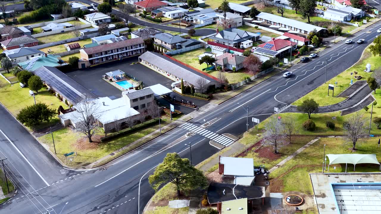 Drone camera glides above quiet suburban neighborhood with residential buildings, swimming pools, and intersecting roads under bright daylight, capturing a peaceful small-town atmosphere