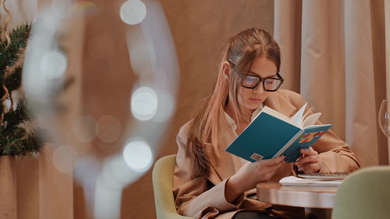 Femme caucasienne lisant un livre bleu, lumière chaude douce et confortable, bokeh au premier plan, lunettes, assise à une petite table avec un rideau en arrière-plan, tournant les pages, expression contemplative, détente en soirée