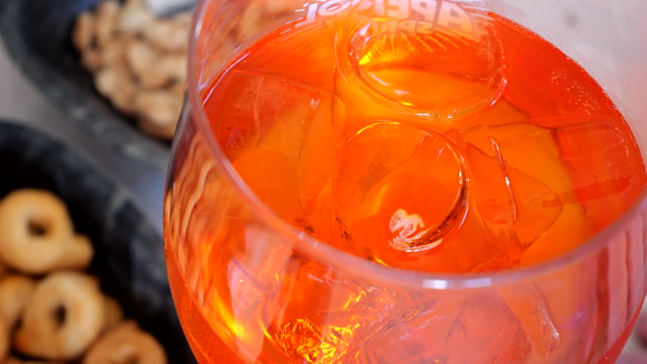 Detailed view of a vibrant orange Aperol Spritz with ice cubes in a stemmed glass, served next to assorted snacks, captured by macro bar scene shot