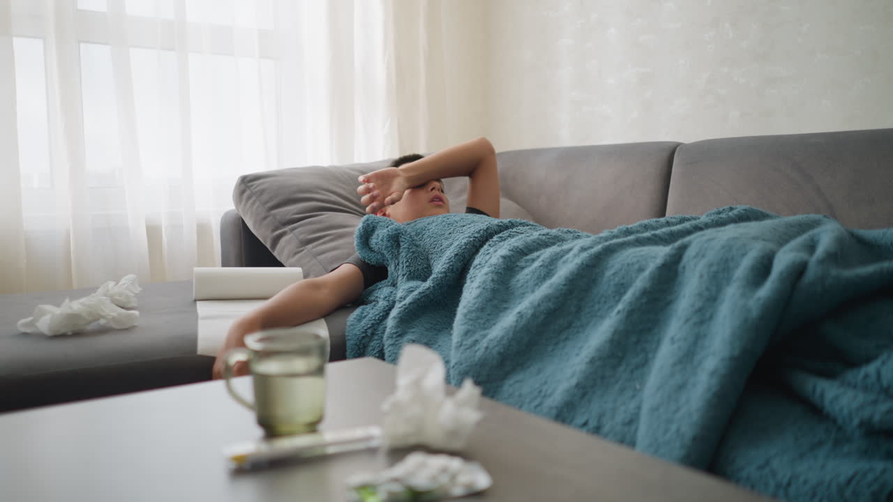 Sick boy resting on couch, hand on forehead, with used tissues, thermometer, and glass cup on the table, boy covered in blanket, recovering from illness in living room