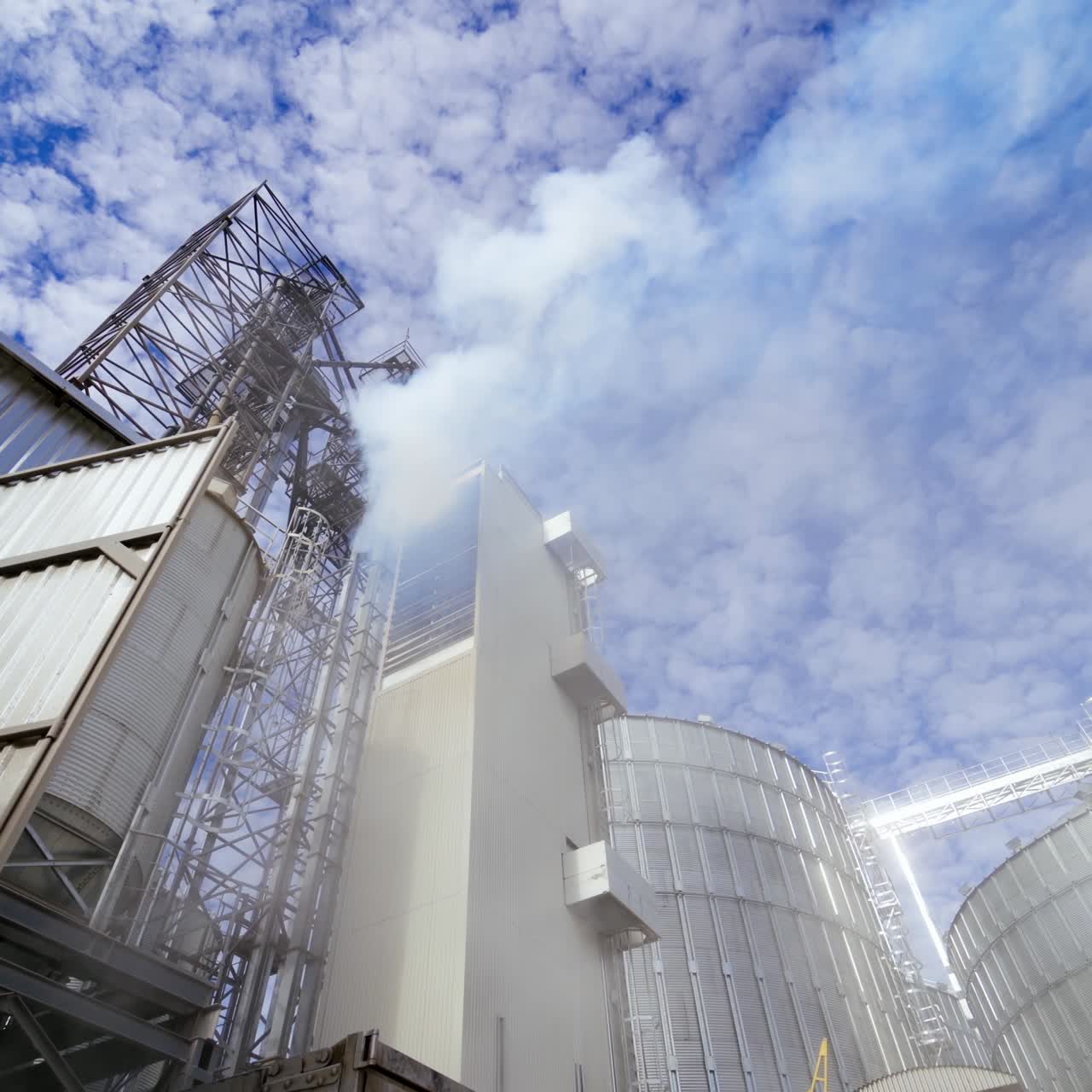 Modern plant with metal elevator for agribusiness. White smoke from industrial grain dryer on sky background. Storage and further processing of grain. View from below.