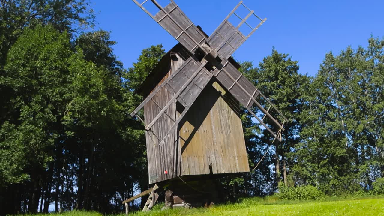 Low angle view of an old traditional wooden windmill at a sunny and grassy countryside by a forest during a summer day. large wooden blades or paddles visible on it that are ready to mill grain