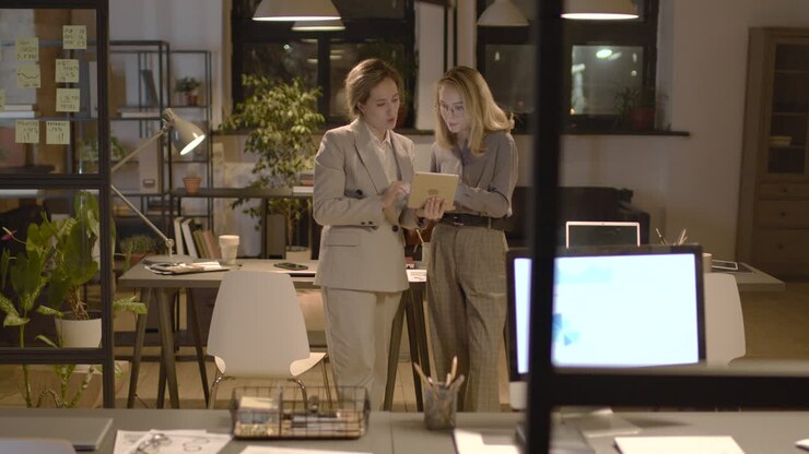 Two Female Coworkers Talking And Watching A Tablet Standing In The Office