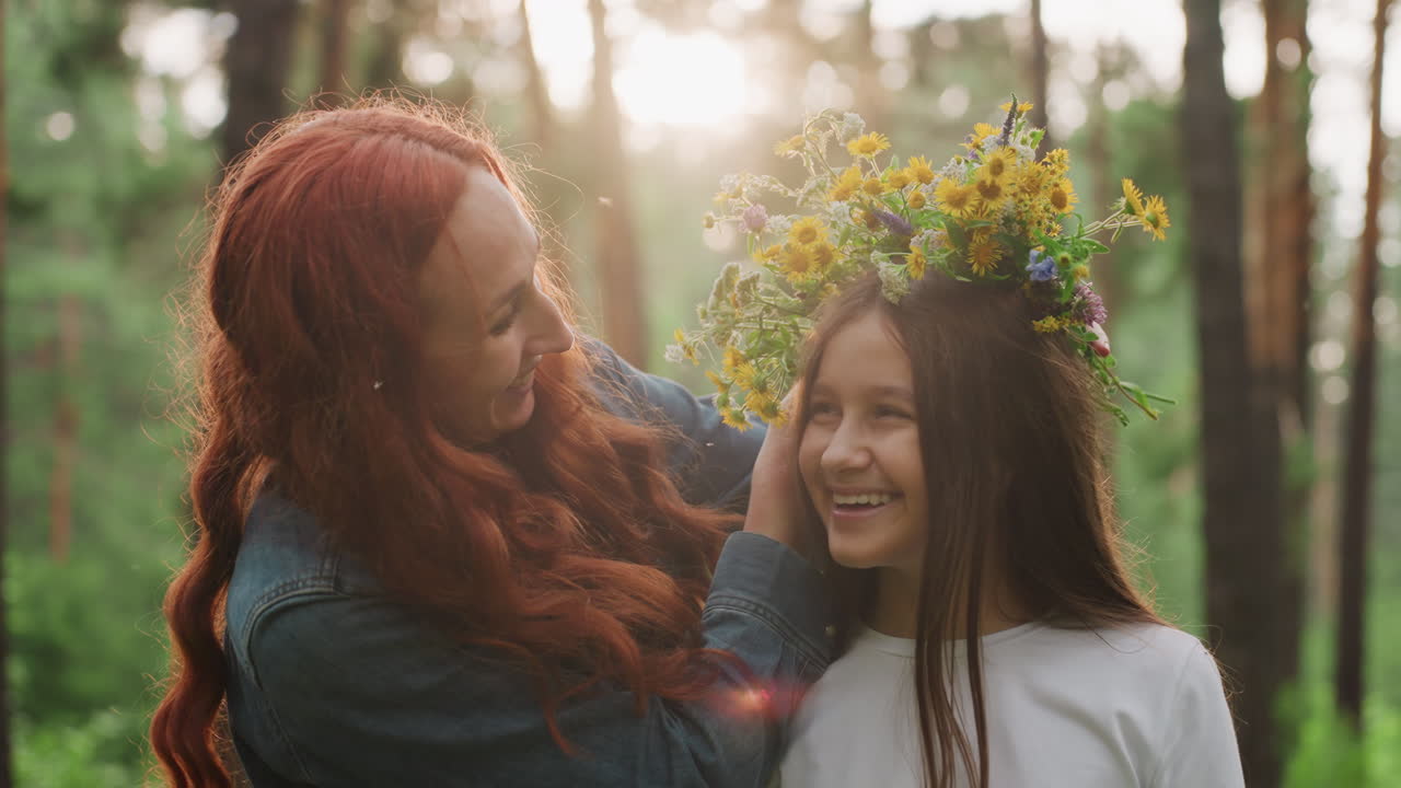 Young mom with red hair decorates her little princess with bright wildflowers in green forest, smiling with tenderness as sunlight shines through trees, capturing warm loving family connection