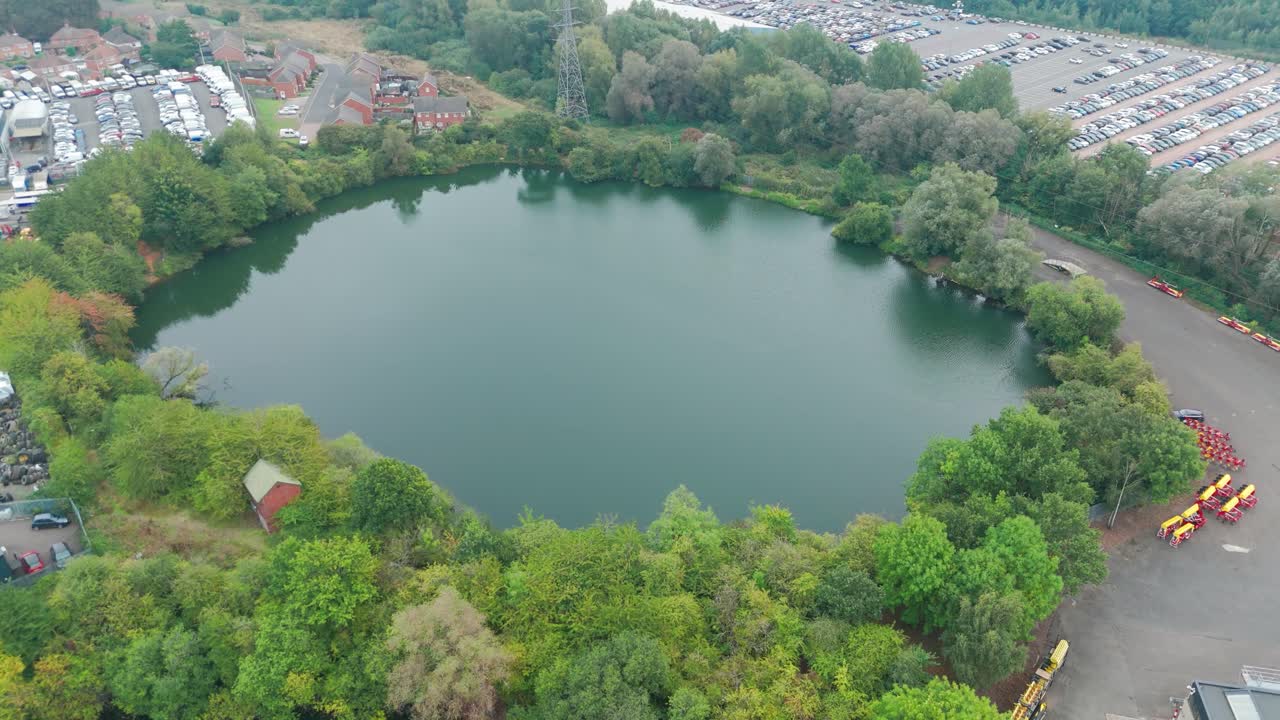 An aerial photo of a circular lake surrounded by trees. The water is dark green and calm. A small building can be seen on the shore. There is a parking lot with cars visible in the background.