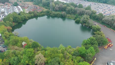 an aerial photo of a circular lake surrounded by trees. the water is dark green and calm. a small building can be seen on the shore. there is a parking lot with cars visible in the background.