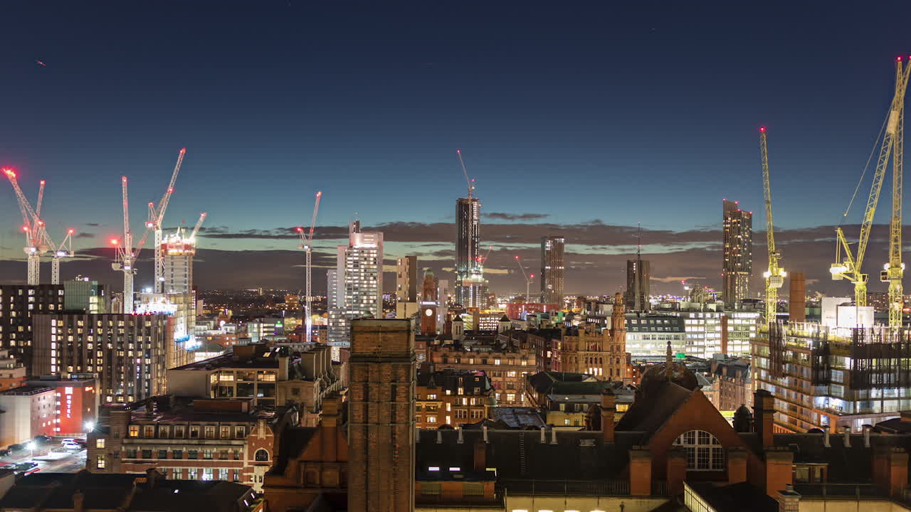 Blue hour timelapse view of Manchester skyline. Clouds move across sky and cranes operating on skyscraper construction projects. Lights of the city at night. Zoom out.