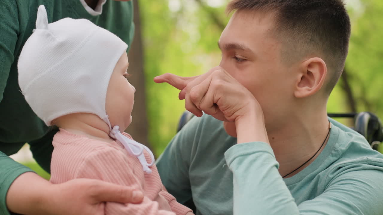 CloseUp White Father Making Funny Faces To Entertain Baby, Playful Interaction Near Wheelchair, Animated Expressions, Tiny Fingers Reaching, Joyful Outdoor Mood, Bonding Through Laughter