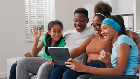 a happy family sits on the couch, relaxing, spending time together in the afternoon, parents with two daughters waving at the tablet camera, having a video conversation with friends