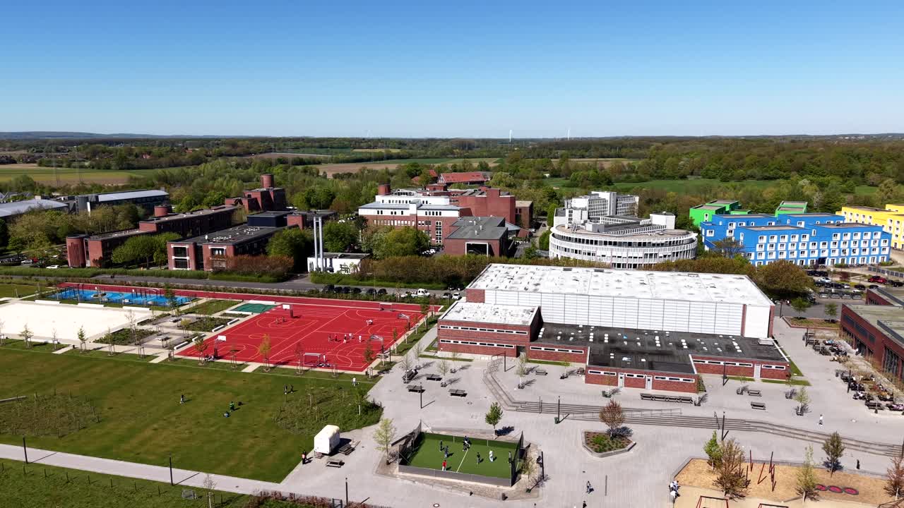 Orbita aerea di un moderno campus scolastico in primavera, con studenti che giocano su campi sportivi rossi, edifici accademici in mattoni, ampi campi verdi e cieli soleggiati in Europa. Campo lungo. Giornata di sole in primavera.