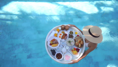 overhead view of a female tourist walking in a crystal clear pool with a floating breakfast with delicious food on it