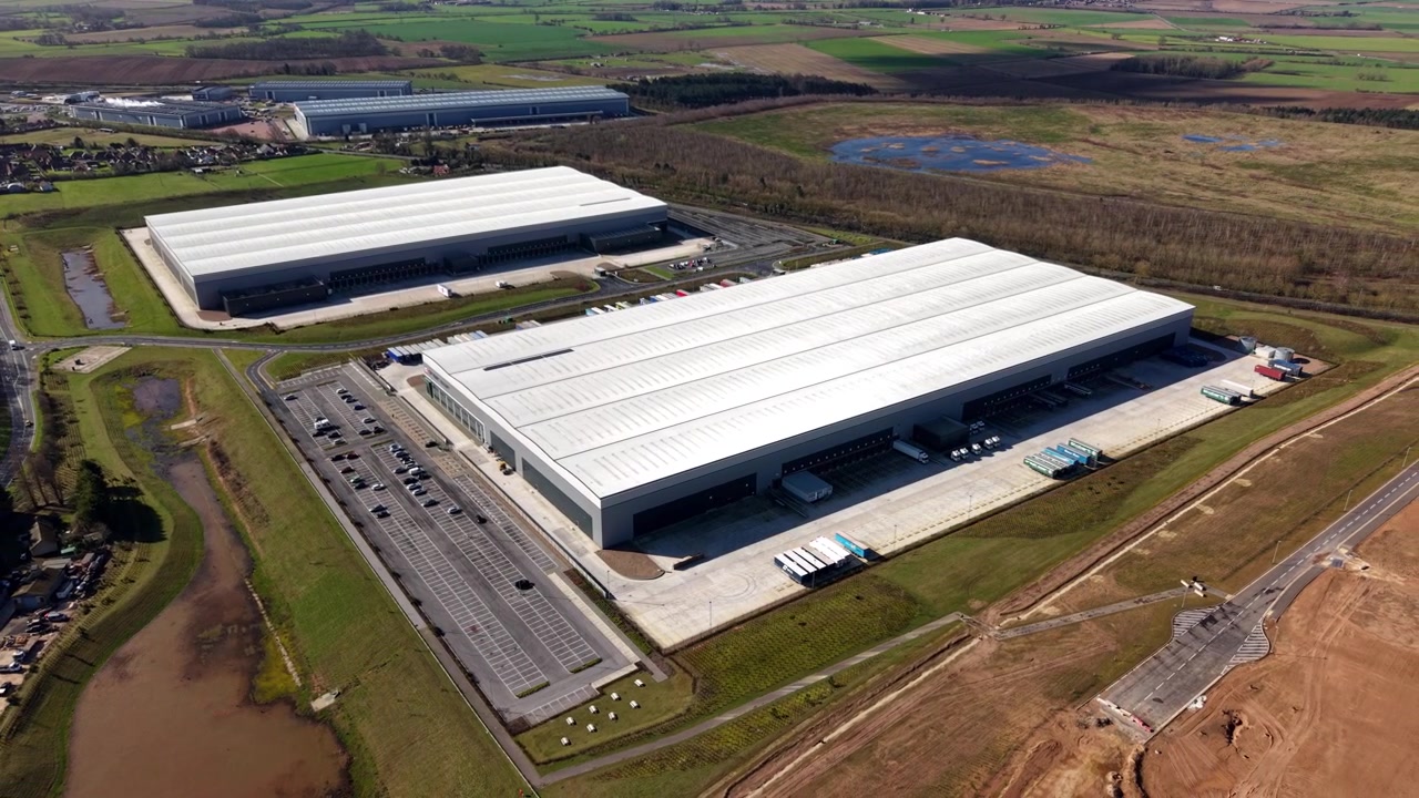 Enorme parco logistico industriale con autocarri, corsie attive per il transito merci e strutture per la spedizione merci, immerso nei campi della campagna rurale britannica
