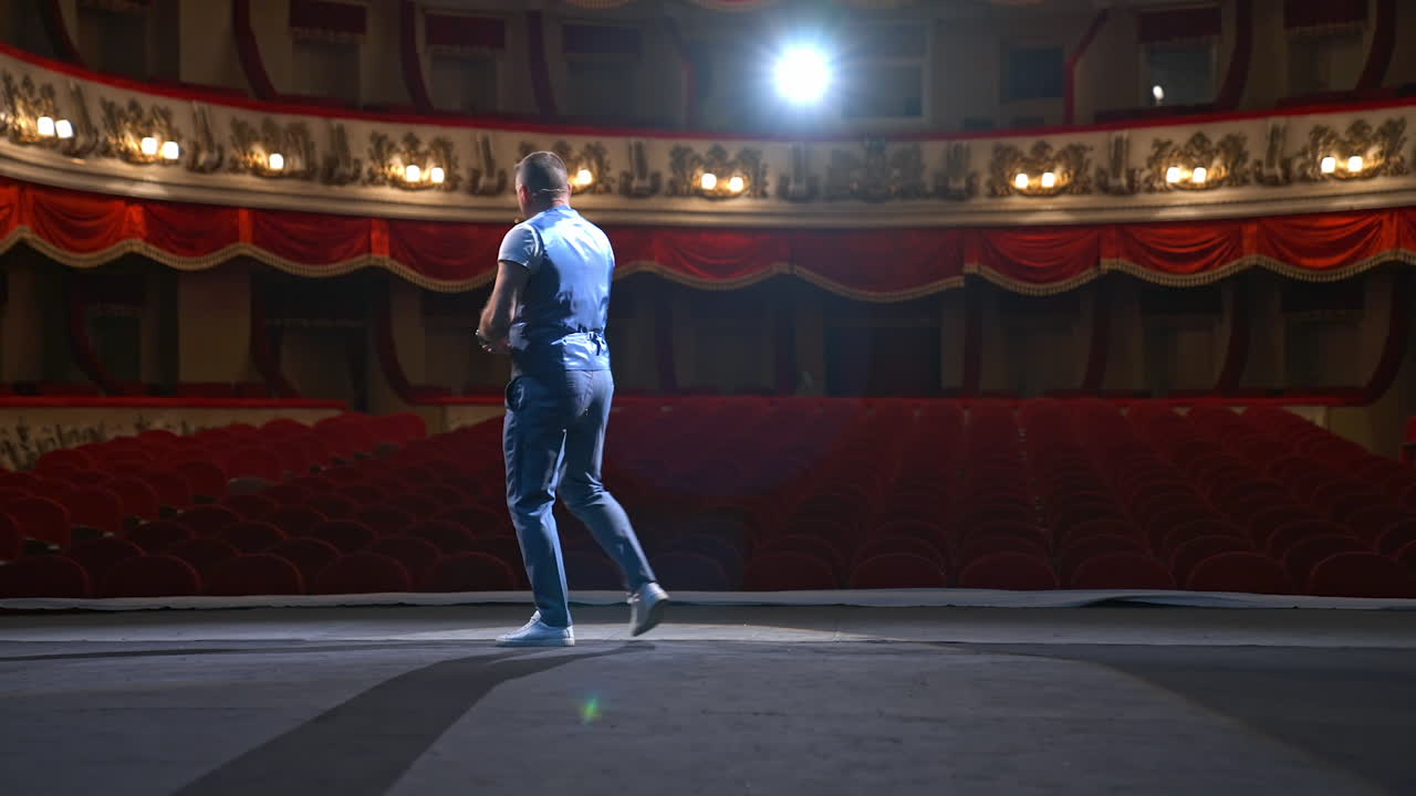 Man in front of empty theatre hall with red velvet chairs. Actor is standing on stage and talking with gestures before the performance.