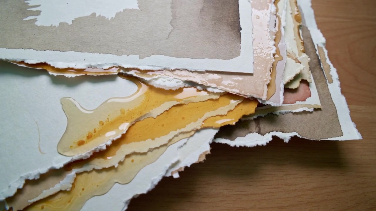 Seeping amber liquid from right edge soaking stacked deckled papers on wood table, darkening washes