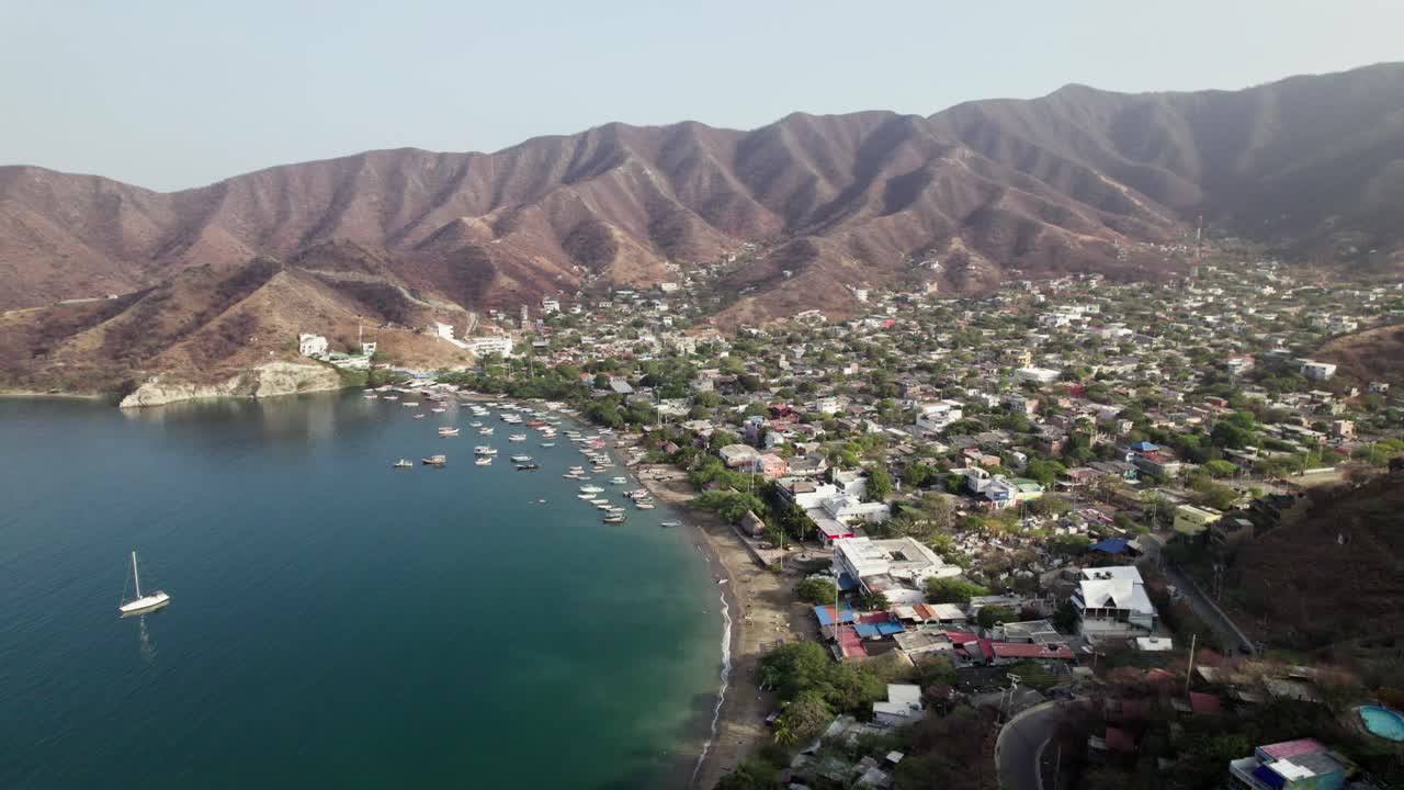 A sweeping drone shot of Taganga, Santa Marta, revealing the picturesque bay, its many colorful fishing boats, a single yacht, and the contrast of the dry, mountainous Sierra Nevada landscape