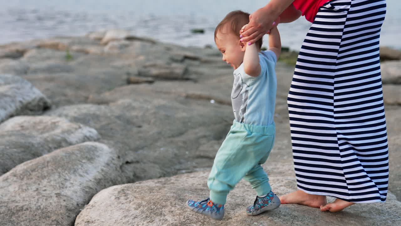 Cute little baby studying to walk at the rocky shore. Mommy is holding her son's hands. Water at backdrop.