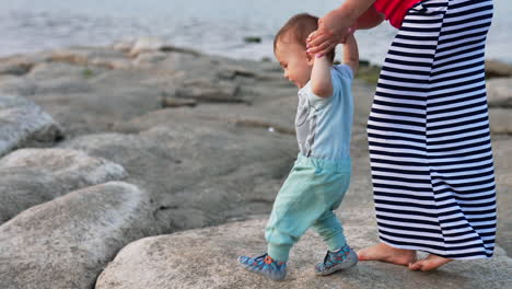 cute little baby studying to walk at the rocky shore. mommy is holding her son's hands. water at backdrop.