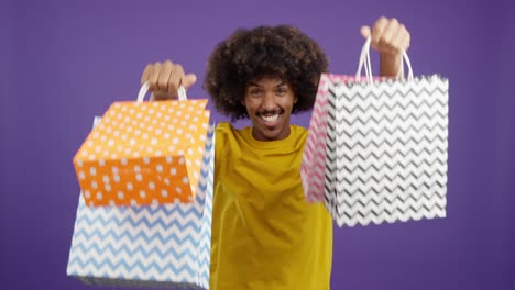 happy man with afro celebrates shopping with bags on purple background