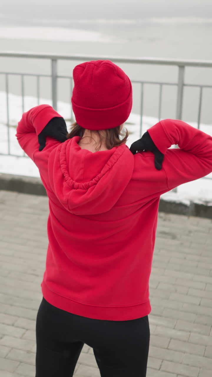 은 후디와 베니를 입은 여성이 어깨에 손을 들고 팔을 바으로 돌리는 팔 스트레칭을 수행하는 뒷면, 배경에는 강과 철 레일링이 있는 안개 분위기