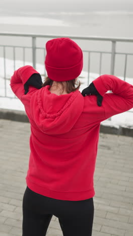 vista trasera de una mujer con capucha roja y gorra realizando estiramientos de brazos levantando las manos a los hombros y girando los brazos al aire libre, atmósfera brumosa con río y barandilla de hierro en el fondo