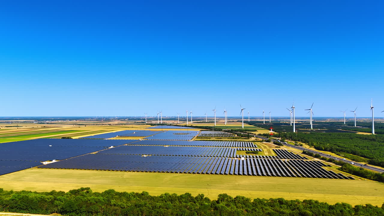 Large sites of modern solar panels in the rural area. Multiple windmills rotate in the wind at backdrop. Aerial view