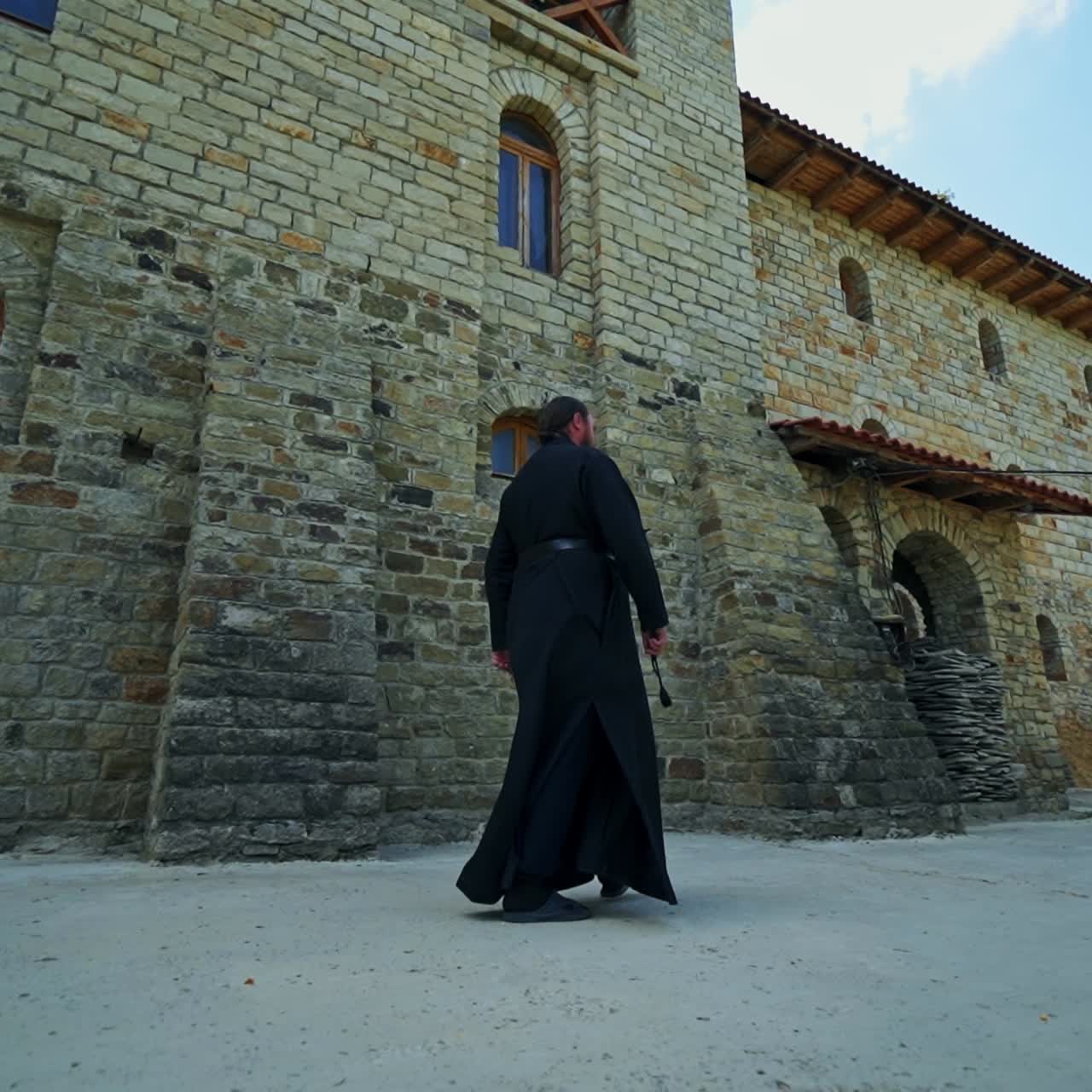 Priest in black clothes walking along the stone walls of monastery. Religious of the orthodox church. Low angle view