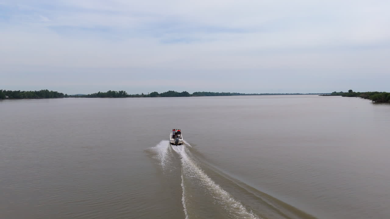 motorboat glides away from the camera through the Paraná Delta’s maze of muddy channels, its white wake fanning out across brown water. Low wooded islets flank the river under a soft gray sky