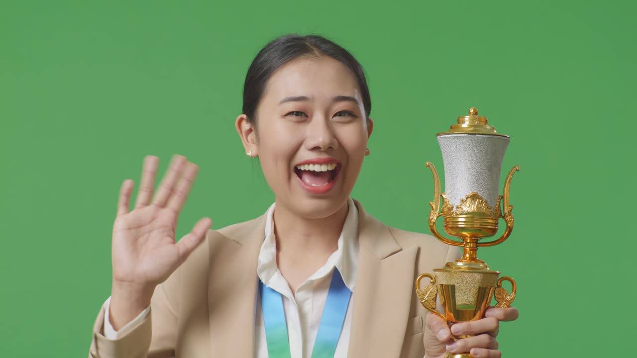 금메달과 트로피를 입은 슈트를 입은 아시아 사업가 여성이 손을 흔들고 카메라에 미소 짓고 스튜디오의 녹색 스크린 배경에서 첫 번째 우승자로