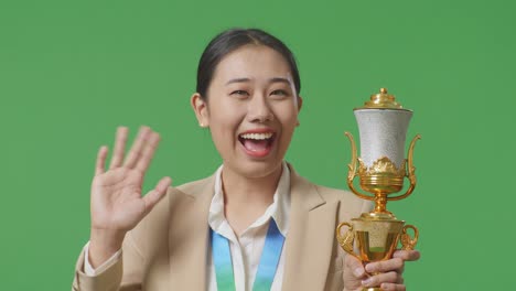 close up of asian business woman in a suit with a gold medal and trophy waving her hand and smiling to camera as the first winner on green screen background in the studio