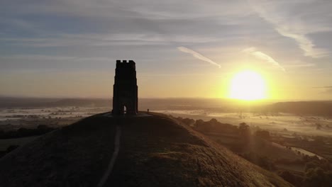 rising aerial of the sunrise overlooking glastonbury tor, revealing the misty fields below