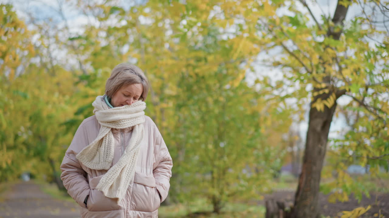 Young woman in light jacket and knitted scarf gently adjusts muffler while strolling through serene autumn park surrounded by colorful foliage
