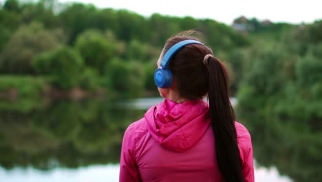 una chica con una chaqueta rosa y auriculares azules se para con la espalda y mira el río temprano en la mañana después de una carrera