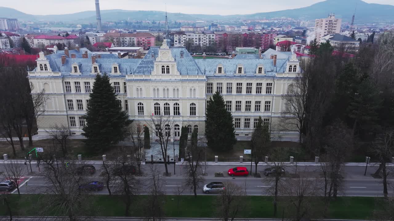 Wide, aerial shot of the historic College "Liviu Rebreanu" in Bistrita, Romania, during the day. The ornate building facade is visible with surrounding cityscape and traffic below