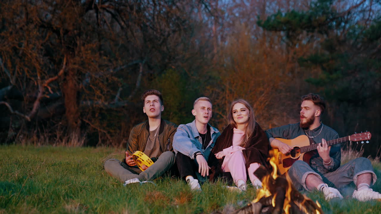 Cosy evening of friends sitting by the fire. Young people singing song together while a man playing the guitar on trees background.