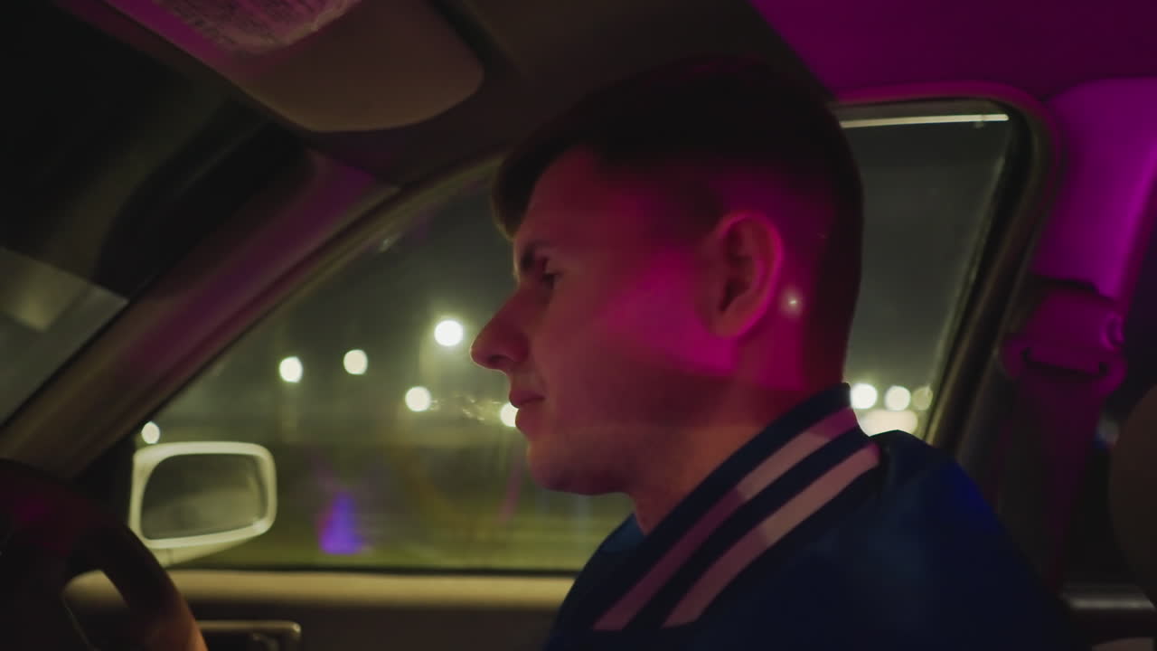 Reflexiones al volante de noche. El interior del vehículo refleja un estado de ánimo contemplativo. Conduciendo por las afueras a un ritmo constante y tranquilo. El conductor, con una chaqueta universitaria, contempla el paisaje urbano iluminado de noche.