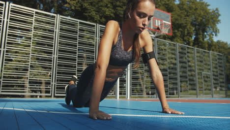 chica de fitness enfocada con airpods haciendo flexiones en la cancha deportiva el día de verano