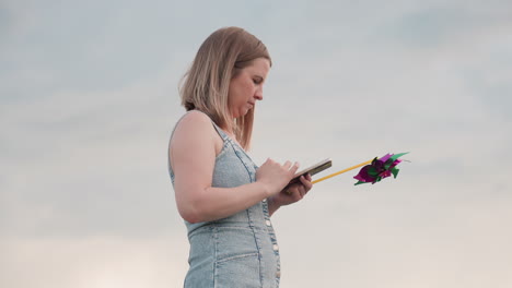 young woman stands on country road holding colorful pinwheel in one hand while operating smartphone with focused expression overlooking green field under serene cloudy sky