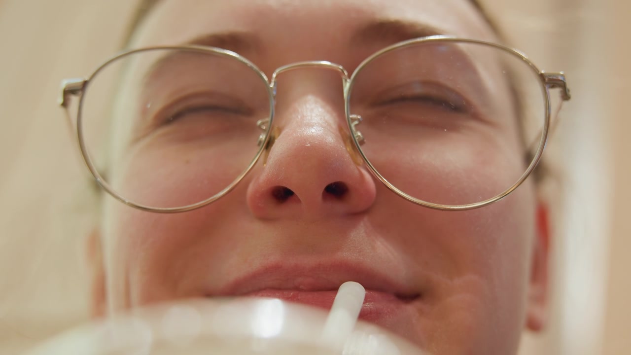 Sguardo frontale con occhiali, sorso giocoso attraverso una cannuccia, giovane che guarda in alto mentre sorseggia una bevanda, montature rotonde e texture della pelle morbida, interno accogliente, scatti di reazione spontanei, risate leggere e quotidianità