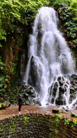 female tourist comes up to picturesque waterfall on the green rock. nature of the azores. vertical video.