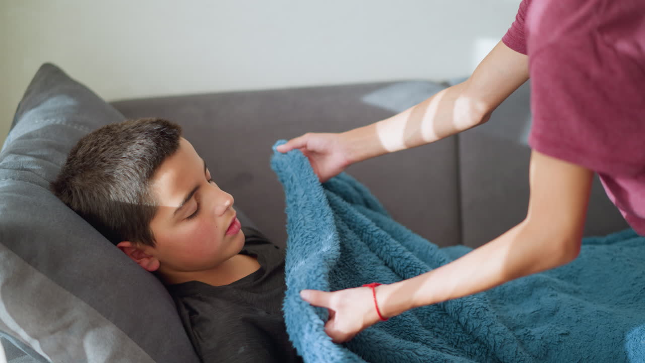 Close-up of sleeping boy on couch with sister coming to cover him with blue blanket, a caring moment as she adjusts blanket to ensure his comfort