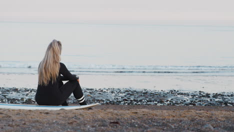 vista trasera de una mujer vestida con traje de neopreno sentada en una tabla de surf y mirando hacia el mar